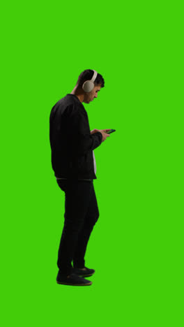 Vertikale-Videoaufnahme-In-Voller-Länge-Eines-Mannes-Mit-Kabellosen-Kopfhörern,-Der-Musik-Vom-Mobiltelefon-Auf-Grünem-Bildschirm-Streamt-3